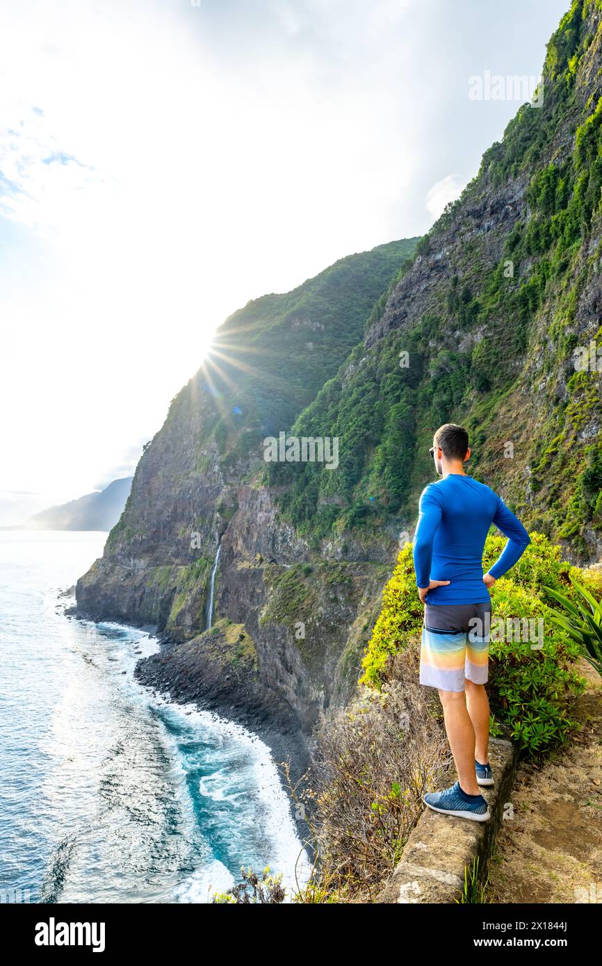 Description: Sporty man looking at waterfall flowing into the sea in atmospheric morning atmosphere. Viewpoint Véu da Noiva, Madeira Island, Portugal, Stock Photo