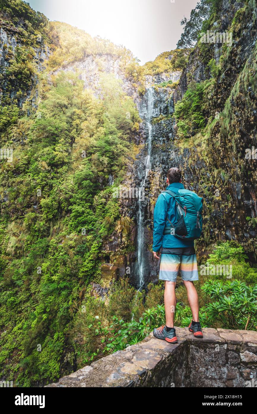 Description: Sporty tourist with backpack standing on stone wall viewpoint and watches green overgrown Risco waterfall. 25 Fontes Waterfalls, Madeira Stock Photo