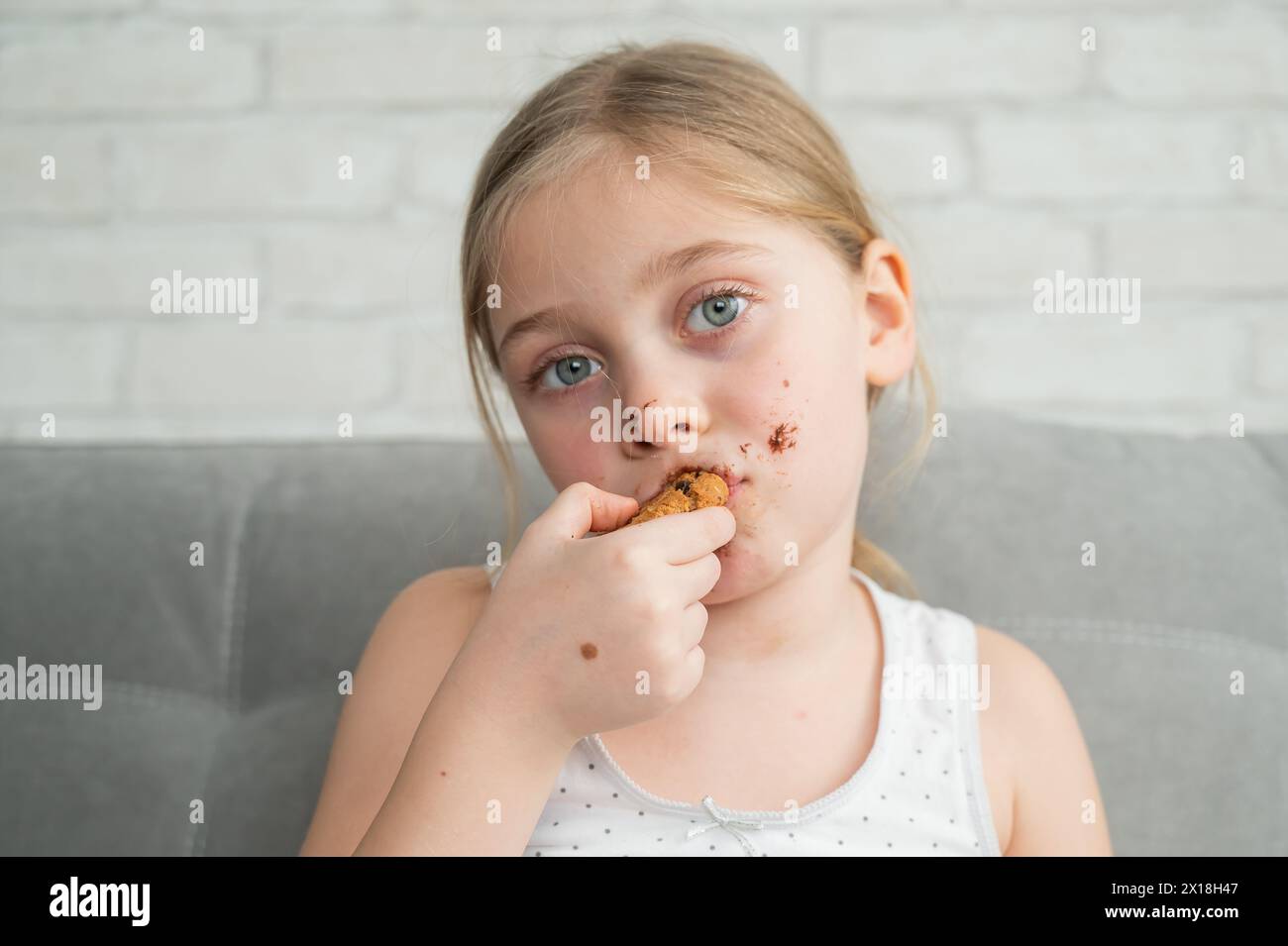 A cute little girl covered in chocolate eats cookies while sitting on the sofa. Stock Photo