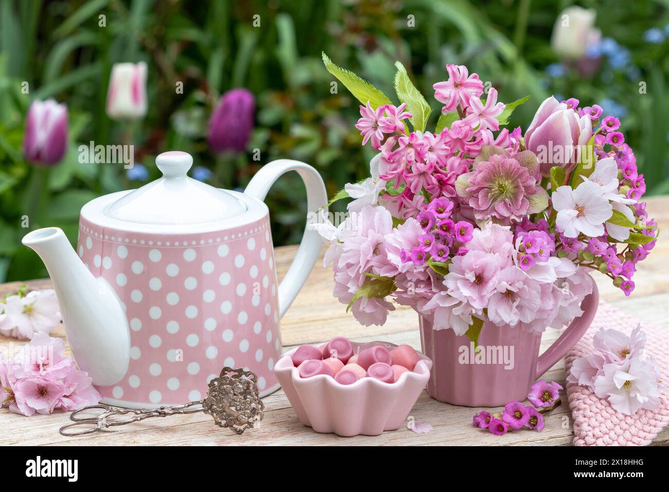 table arrangement with bouquet of lenten rose, tulip, hyacinths, bergenia and Japanese flowering cherry, tea can and candies Stock Photo