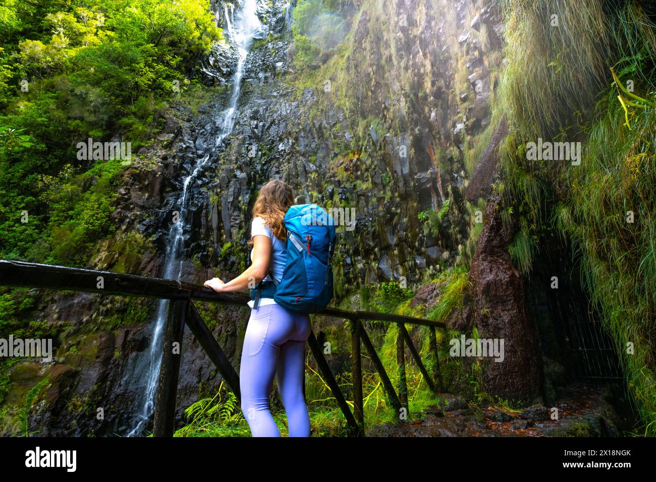 Description: Woman tourist with backpack leans against wooden fence of viewpoint and watches green overgrown Risco waterfall. 25 Fontes Waterfalls, Ma Stock Photo