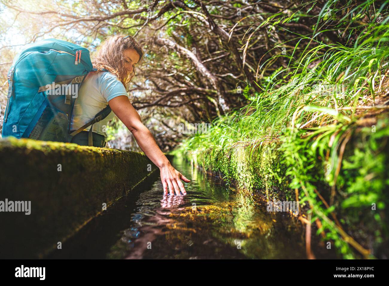 Description: Low angle shot of tourist with backpack holding her hand in fresh water canal along green overgrown hike path. 25 Fontes waterfalls, Made Stock Photo