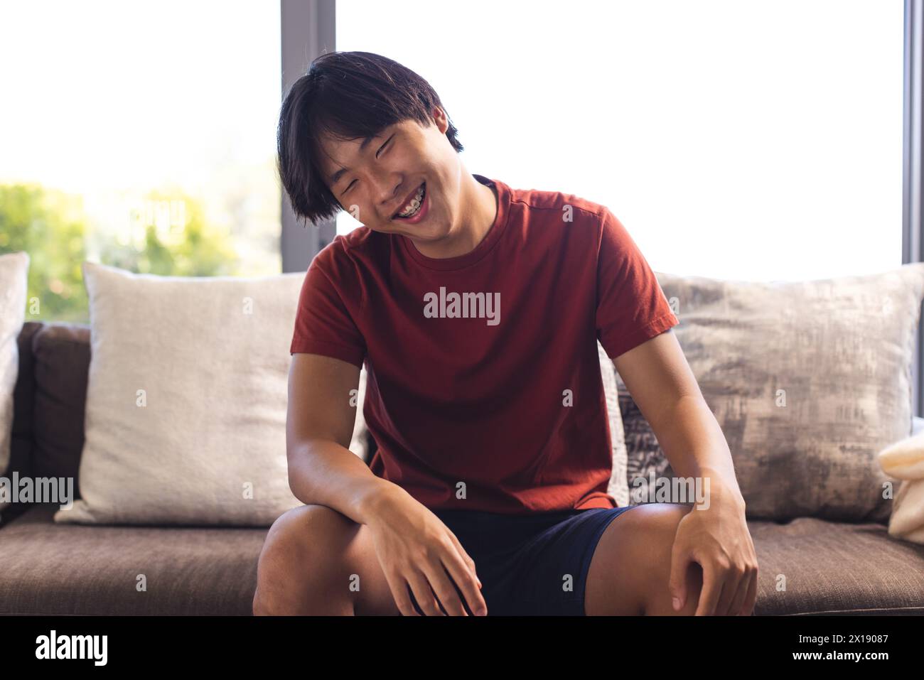 Asian teenage boy sitting on a couch at home, laughing during a video call Stock Photo