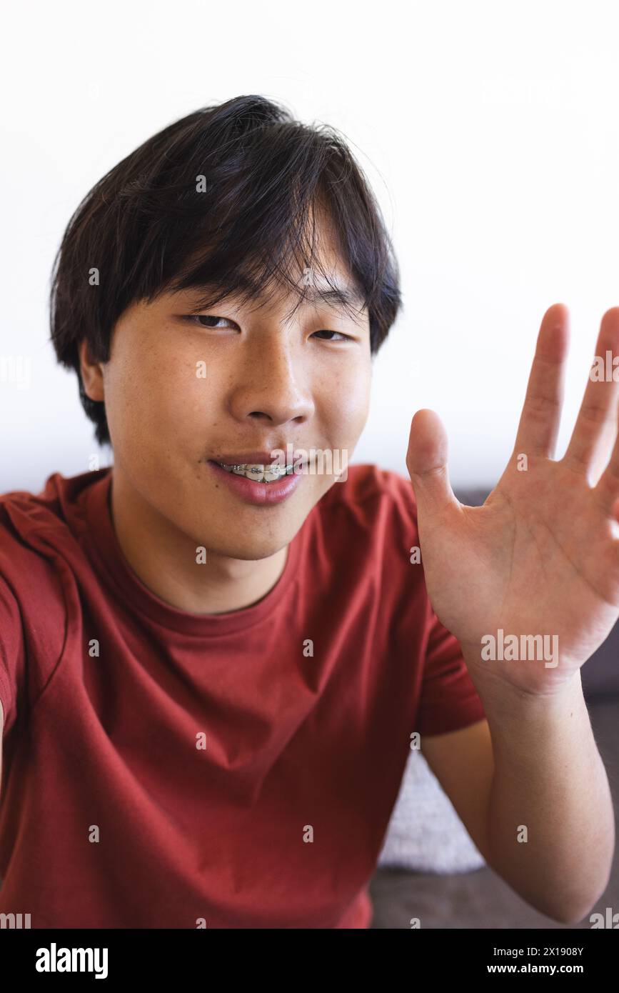 Asian teenage boy waving during a video call at home, sitting indoors Stock Photo