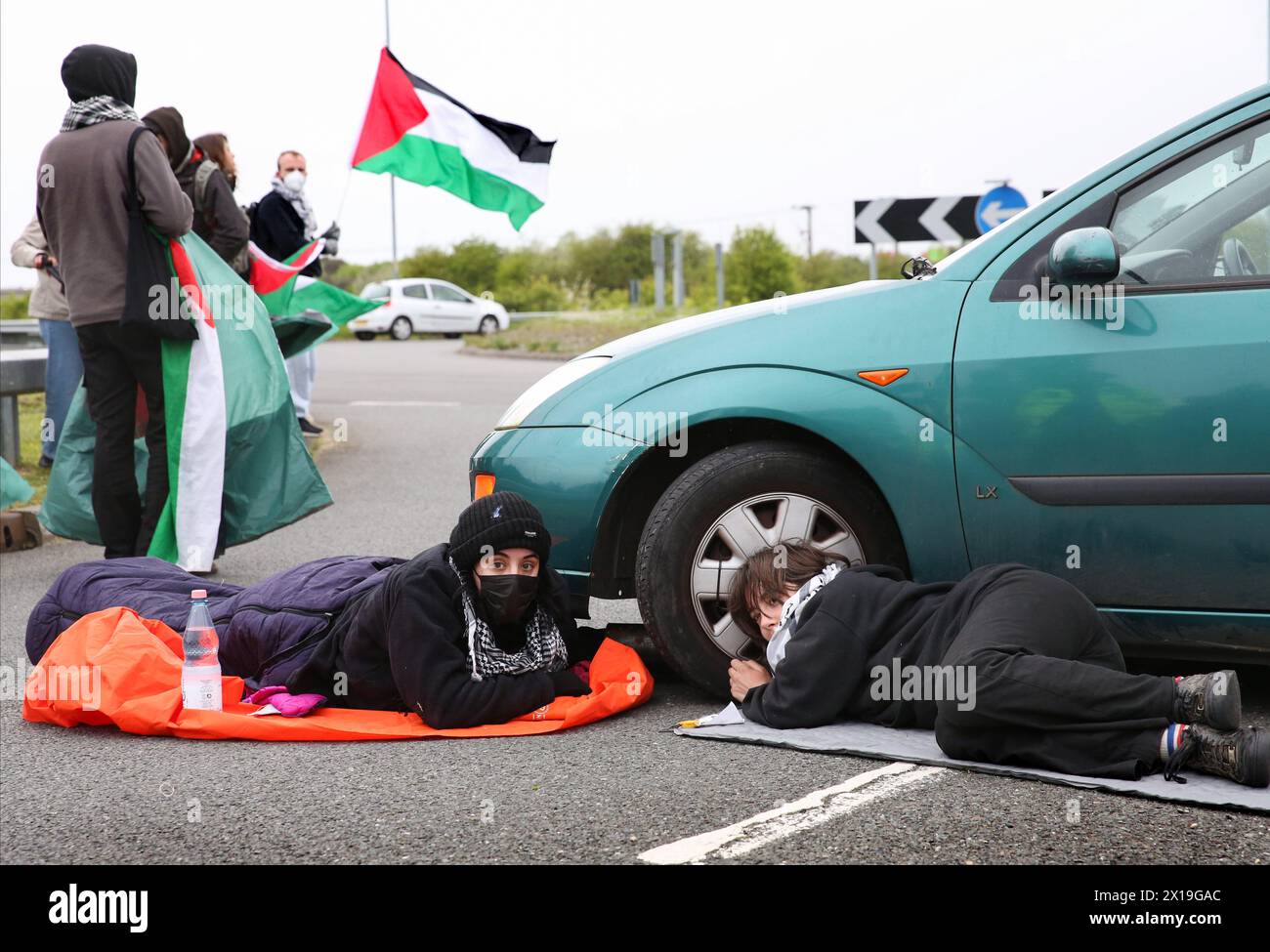 Sandwich, UK. 15th Apr, 2024. Activists from Palestine Action lie in the middle of an access road to Discovery Park locked-on around the wheel of a car. Activists blocked the three access roads to business estate Discovery Park. Instro Precision a company owned by Israeli arms company Elbit Systems has a factory here, where they make sights for Israeli sniper rifles. Protesters argue that these weapons are being used against Palestinians in Gaza and elsewhere. The action was part of the A15 coordinated global economics blockade for Palestine. Credit: SOPA Images Limited/Alamy Live News Stock Photo