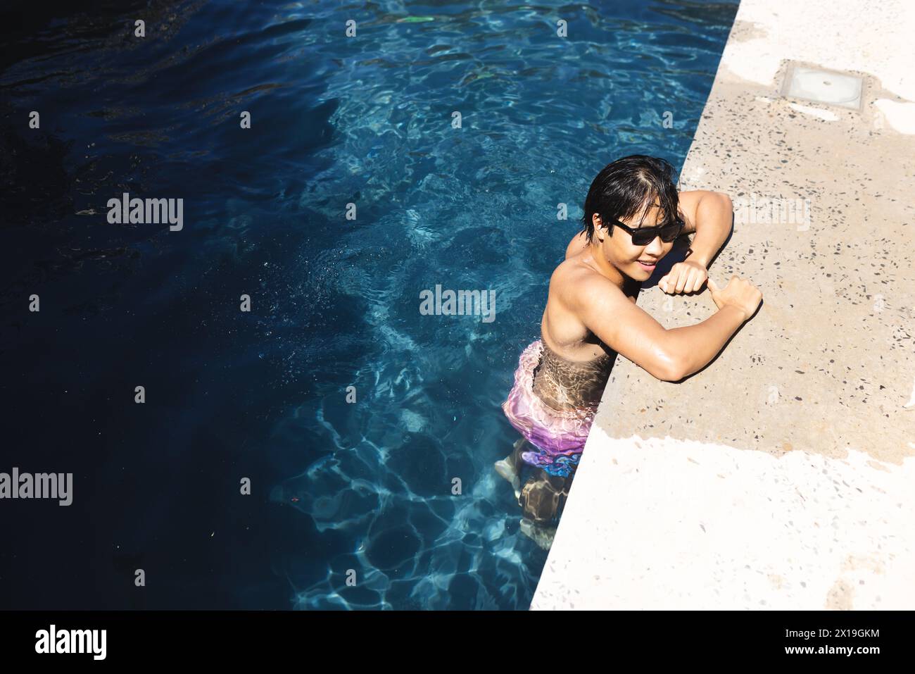 Teenage Asian boy leaning on pool edge outside at home, enjoying the sun, copy space Stock Photo