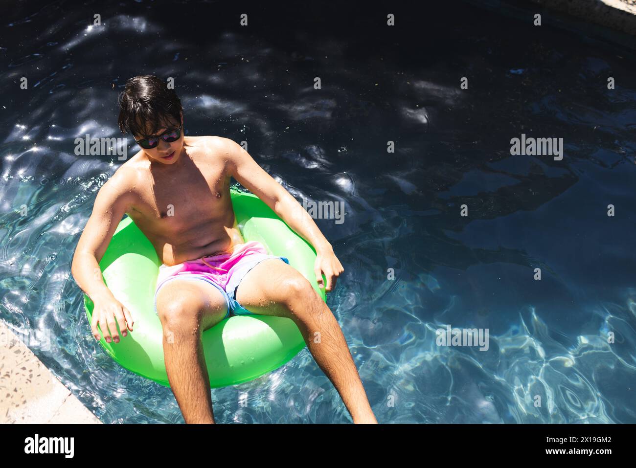 Teenage Asian boy relaxing on a green float in a pool outside at home, copy space Stock Photo