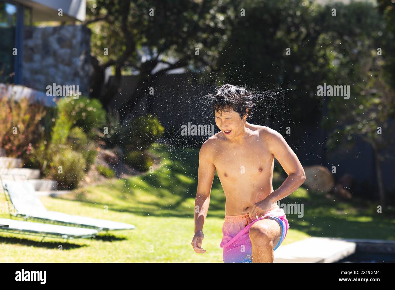 Asian teenage boy running through sprinklers outside at home, enjoying summer Stock Photo