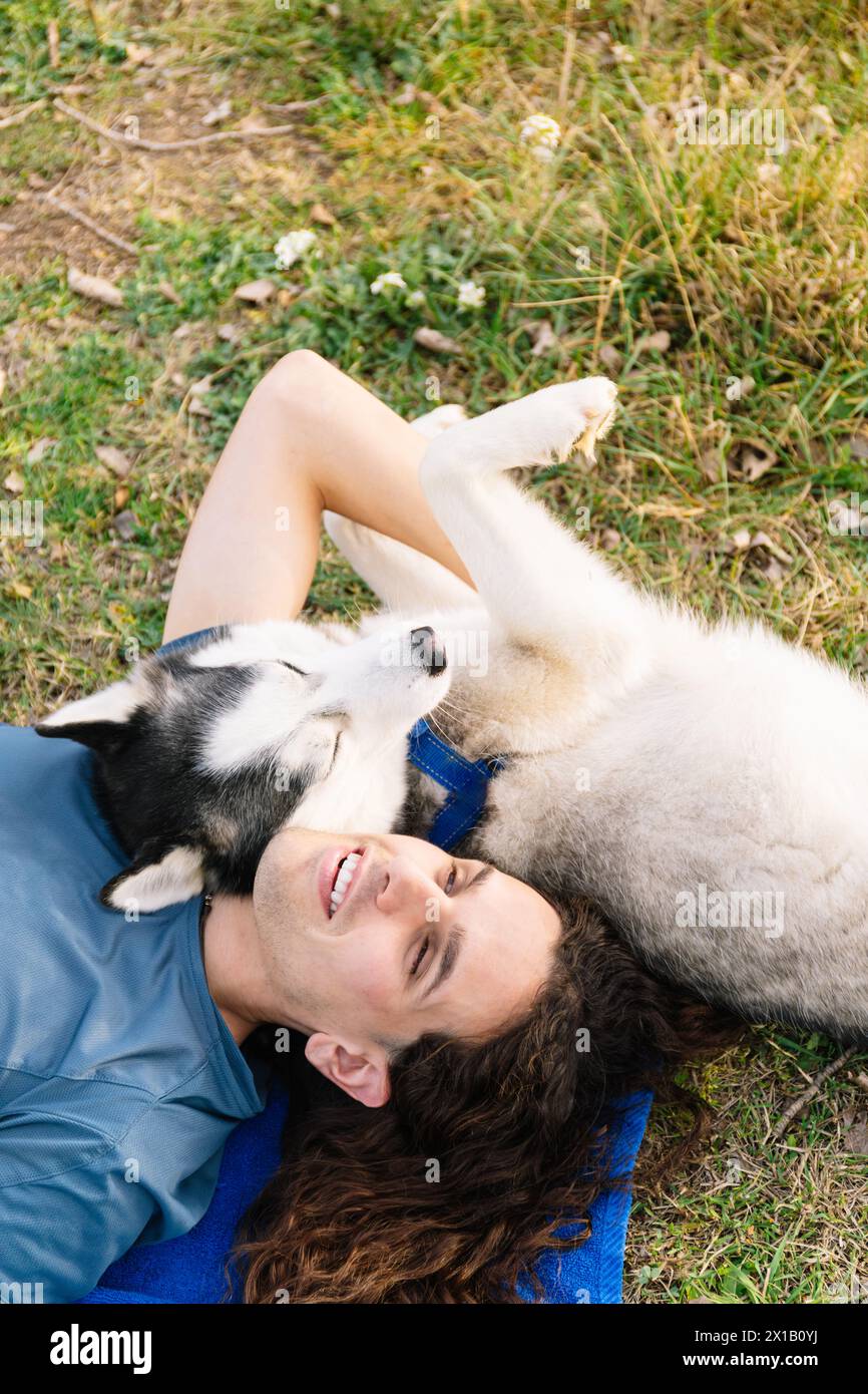 Vertical photo laughter fills the air as a man with curly hair and his playful husky share a moment of pure bliss, lying on the grass togeth Stock Photo