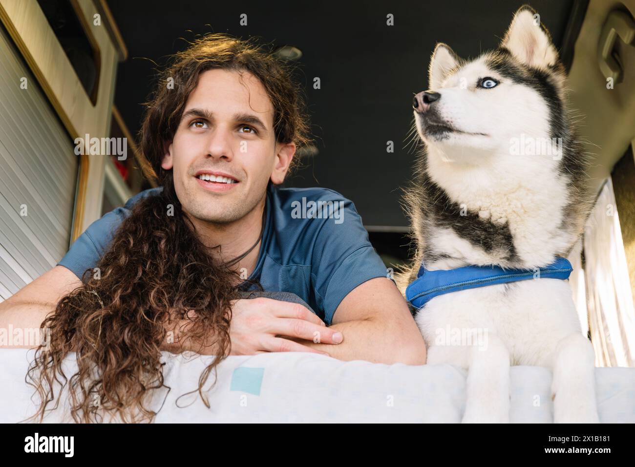 Horizontal photo a relaxed man with curly hair and his blue-eyed husky comfortably lounge inside a camper van, enjoying a moment's rest duri Stock Photo