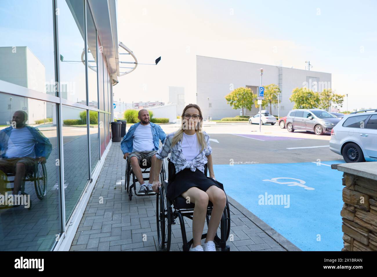 An image capturing two people in wheelchairs, a man and woman, outside a building on a sunny day, showcasing accessibility and mobility. Stock Photo