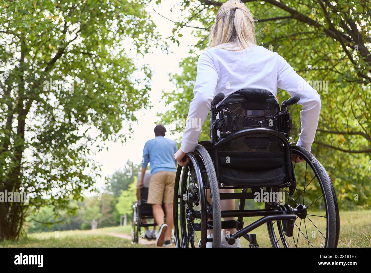 Two individuals who use wheelchairs are spending time outdoors surrounded by greenery, signifying inclusion and accessibility Stock Photo