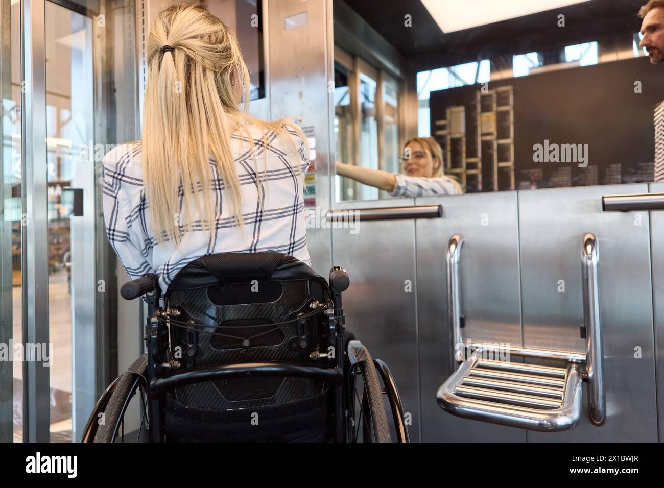 Back view of a person in a wheelchair at a service counter interacting with an employee, showcasing inclusion and accessibility. Stock Photo