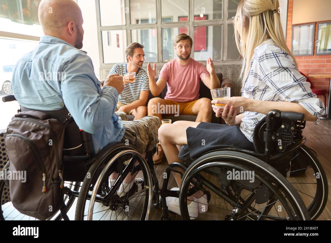 A group of friends, including people who use wheelchairs, engaged in lively conversation and enjoying drinks together in a sunny café setting. Stock Photo