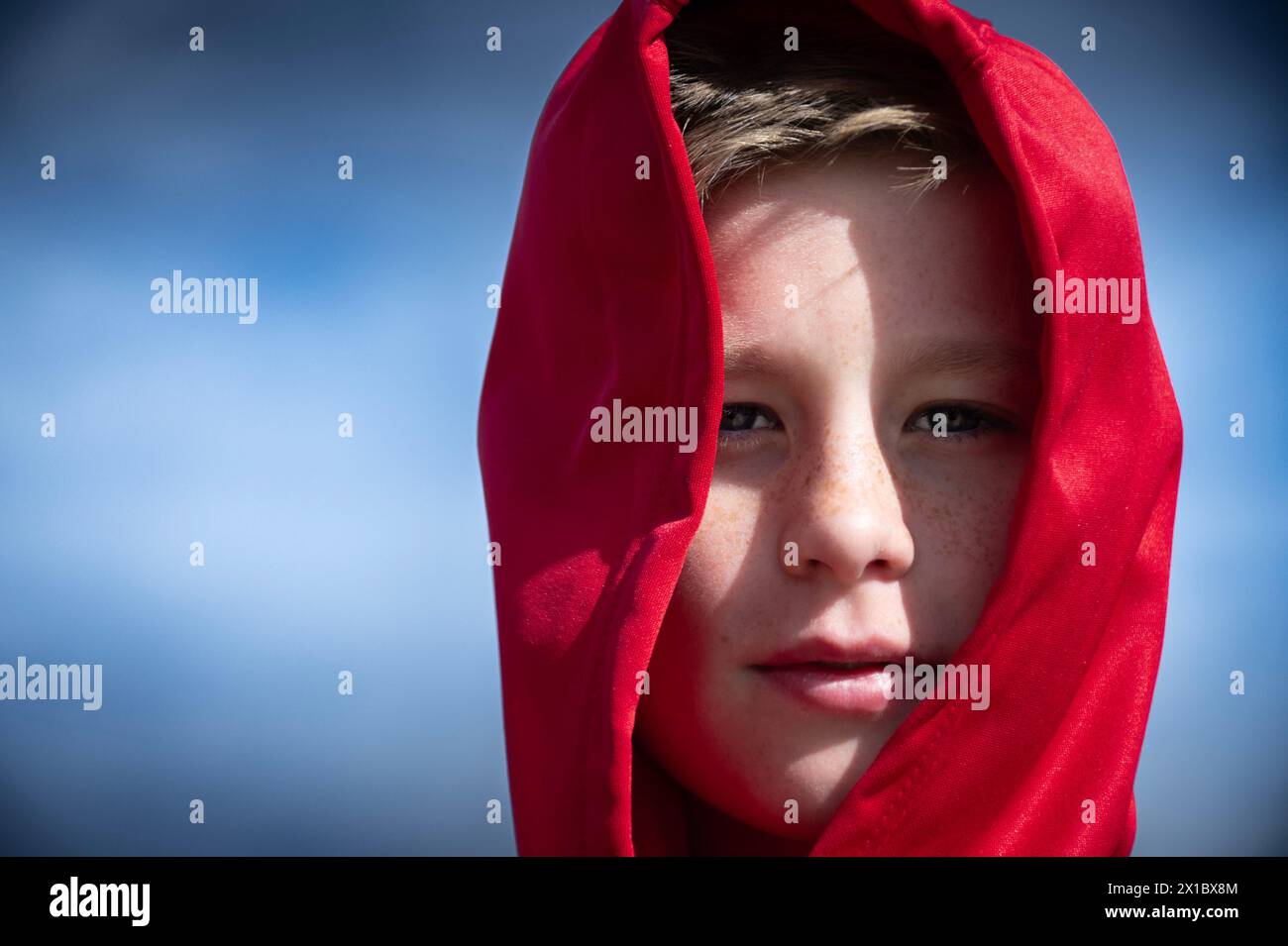 A young boy with light skin and freckles is partially draped in a bright red hoodie that covers his head, leaving his face visible. the background is Stock Photo