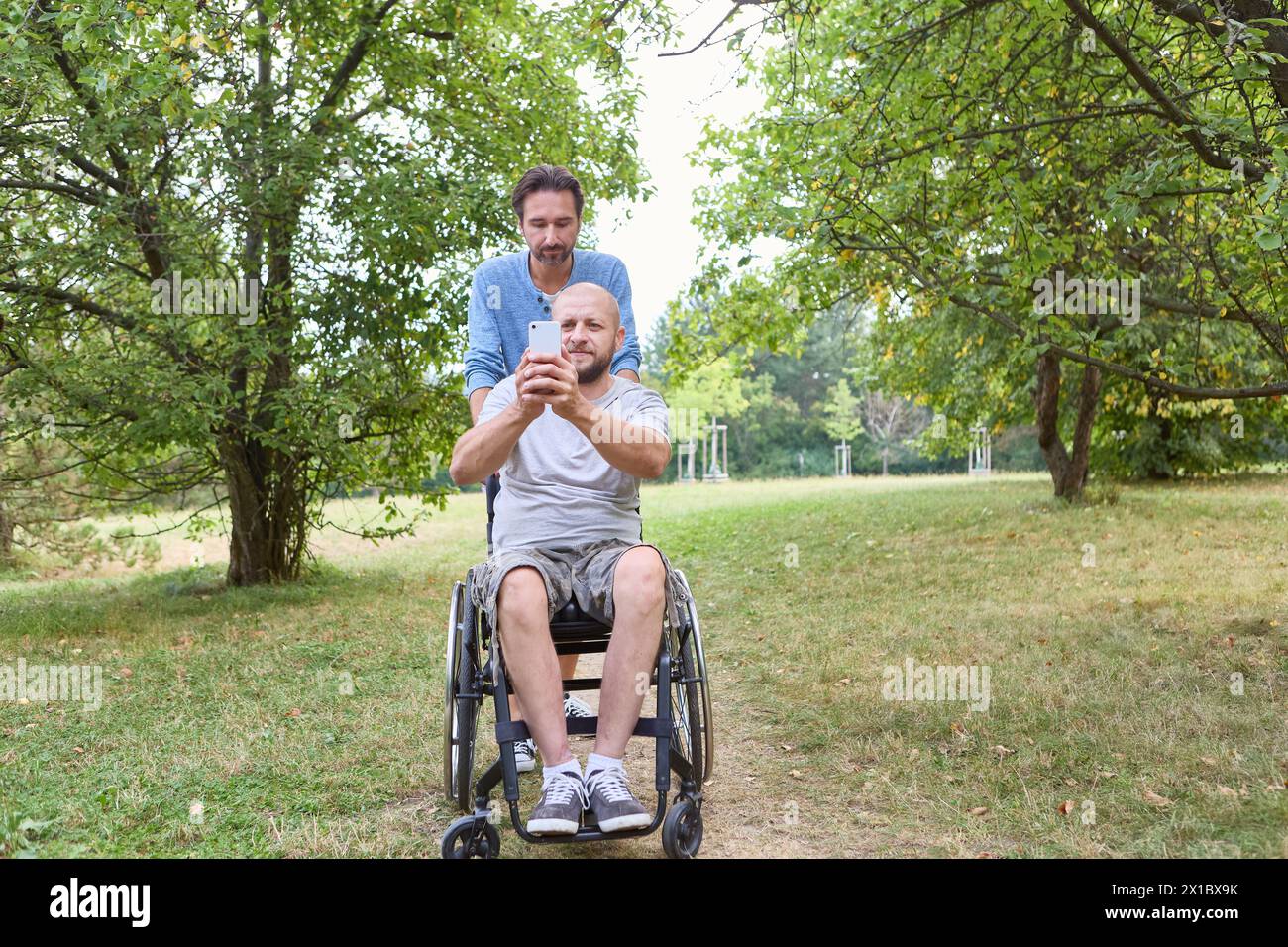 A person who uses a wheelchair and companion share a moment, taking a selfie together in a serene park setting. Stock Photo