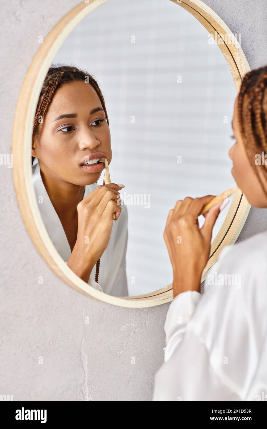 An African American woman with afro braids in a bathrobe brushing her teeth in a modern bathroom mirror. Stock Photo