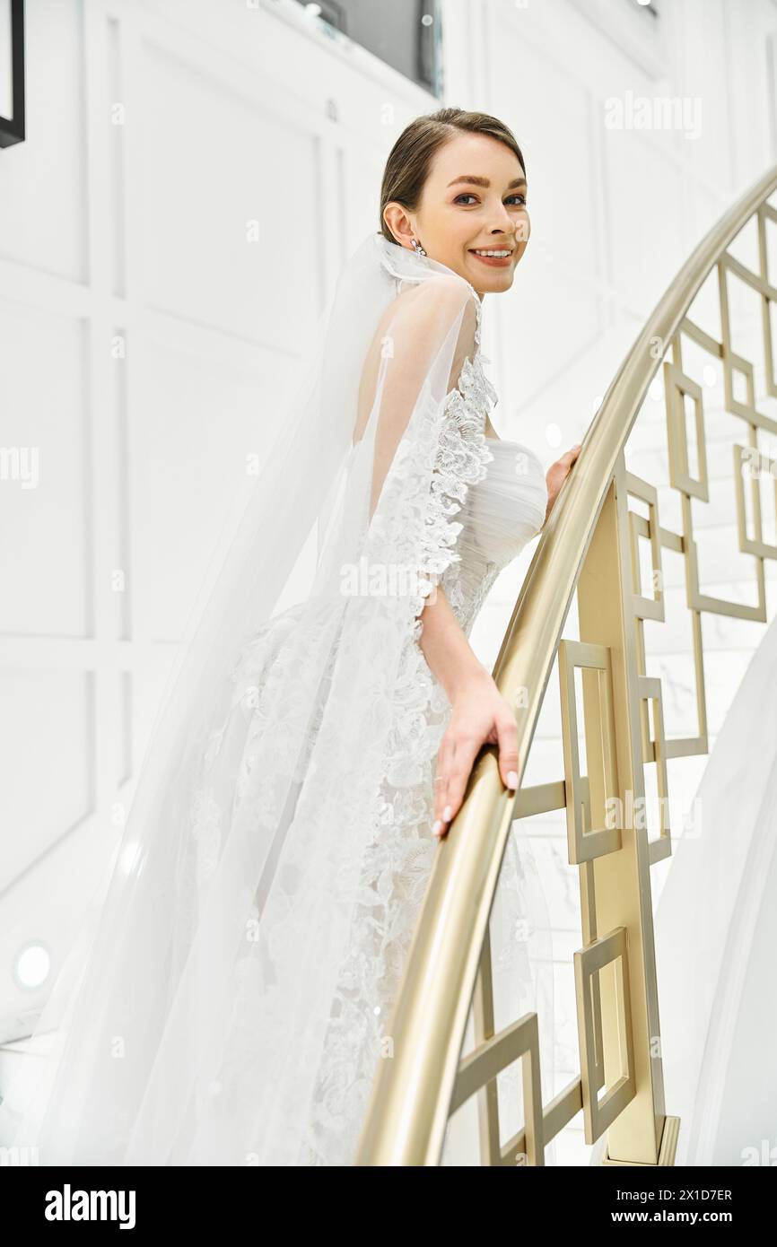 A young brunette bride in a wedding dress stands gracefully on a staircase in a bridal salon. Stock Photo