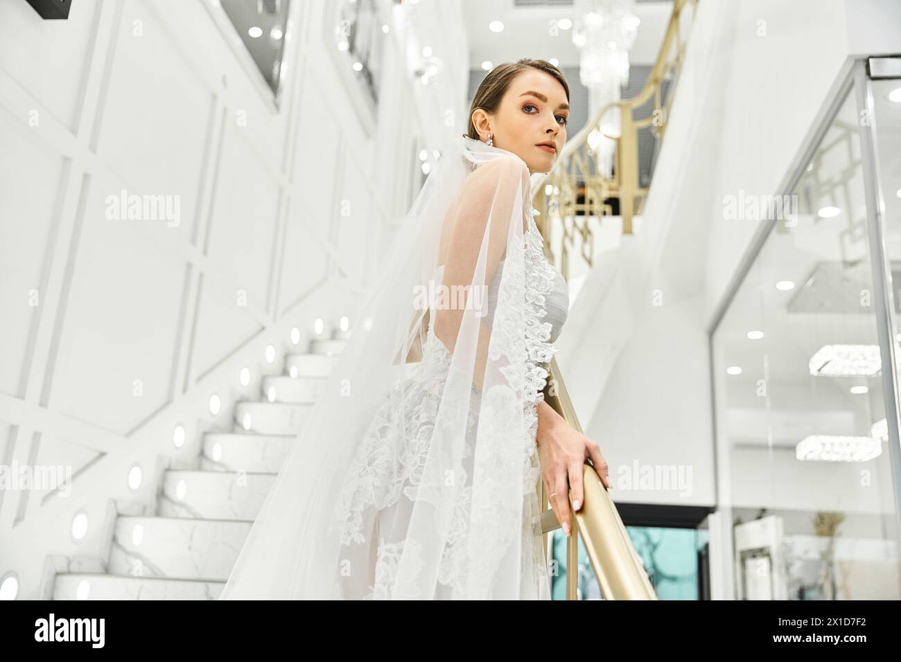 A young brunette bride, dressed in a wedding gown, standing gracefully on a staircase in a bridal salon. Stock Photo