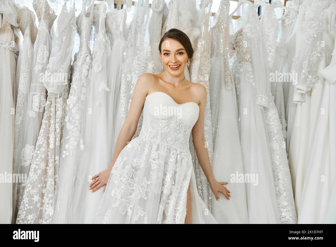 A young brunette bride stands surrounded by a rack of dresses in a wedding salon, looking for her perfect gown. Stock Photo
