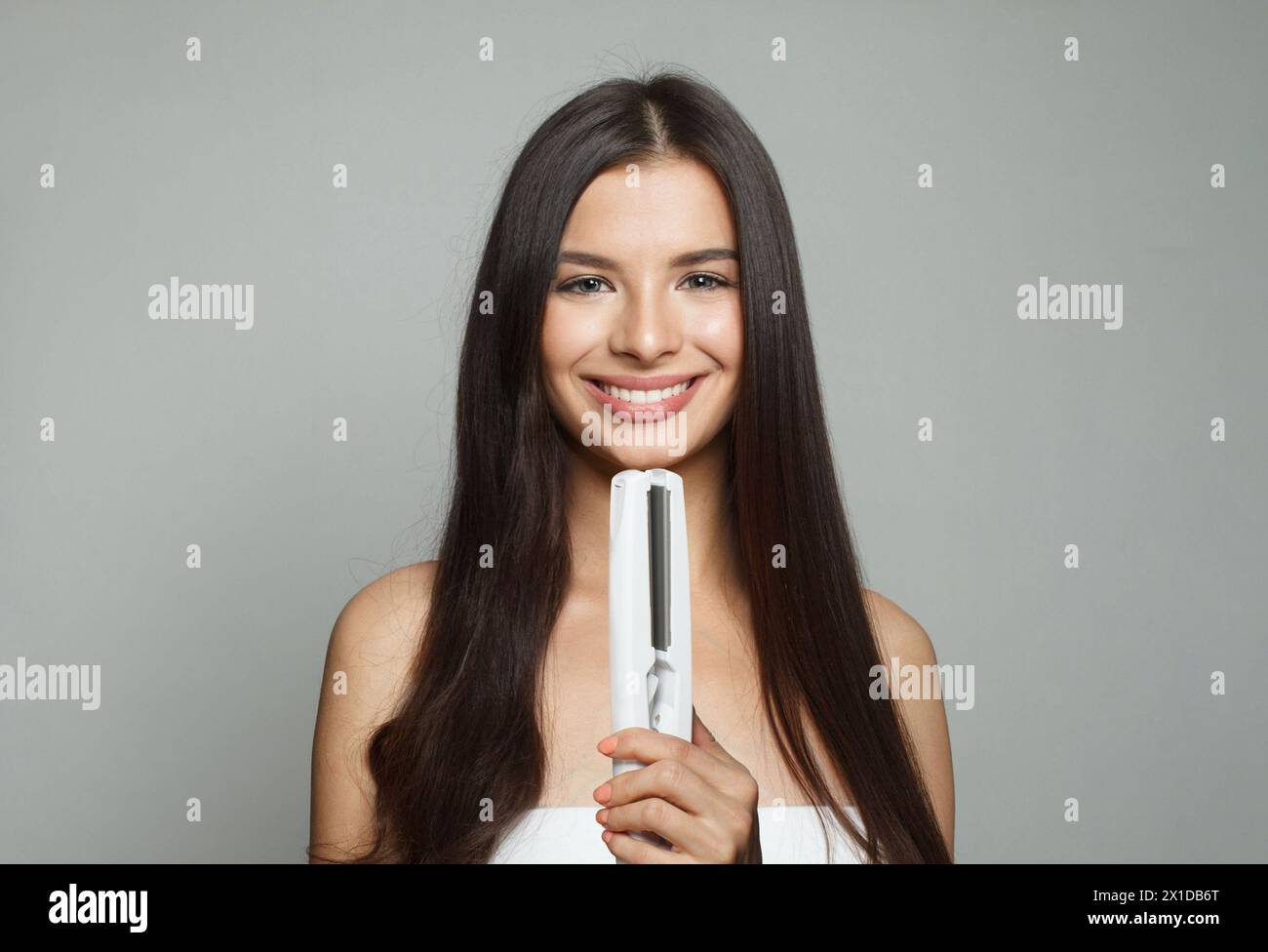 Attractive young healthy brunette woman holding hair iron and straightening her long smooth shiny hair on white background. Haircare, hairstyle, haird Stock Photo