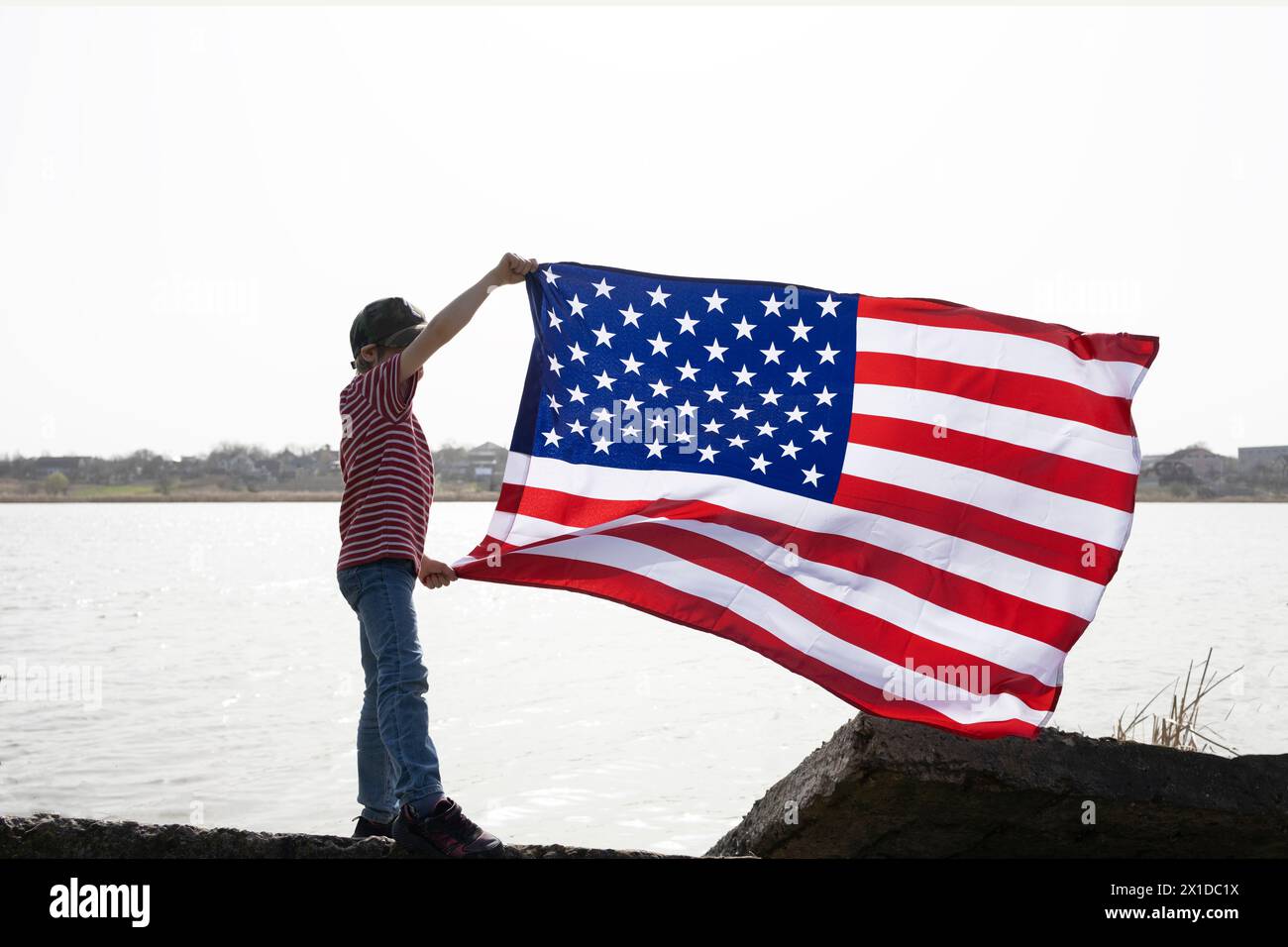 unrecognizable boy stands near lake and holds large waving American flag in his hands. Independence Day. National symbol of freedom of the States of A Stock Photo