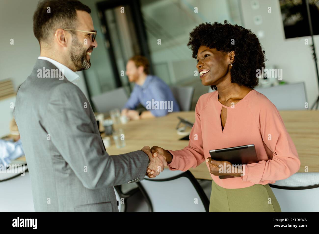 Two professionals warmly shaking hands, smiling in agreement, as teamwork unfolds in the background Stock Photo