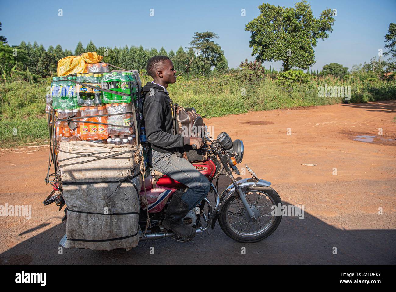A young Ugandan man rides his motorcycle along a dusty, unpaved road, carrying a heavy load of assorted beverages under the warm, sunny sky Stock Photo