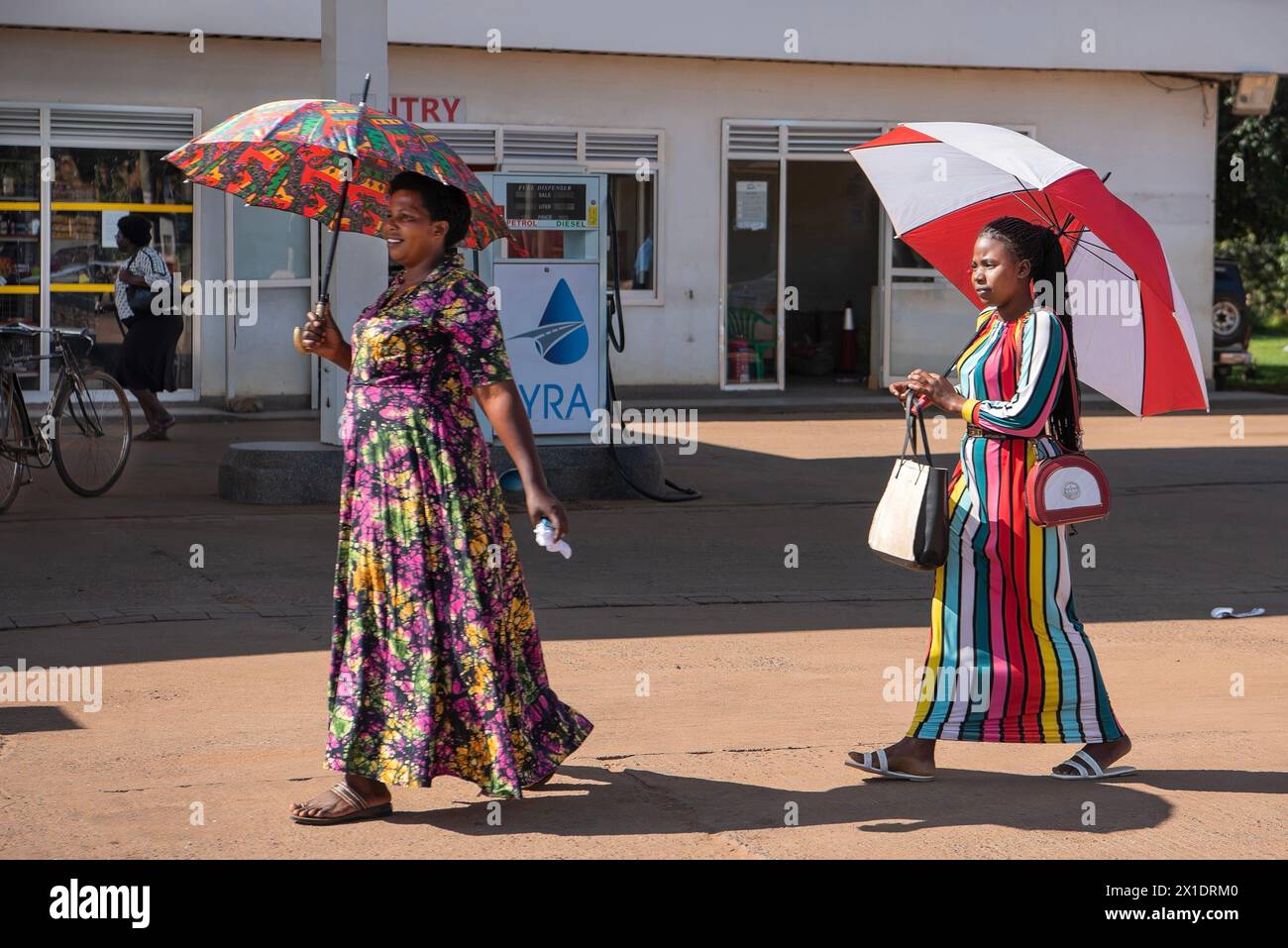 A mother and her daughter, adorned in vibrant dresses, walk gracefully under coordinated umbrellas. They traverse the outskirts of Kampala, Uganda, pa Stock Photo