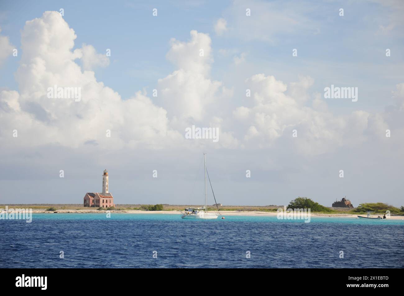 A small sailboat is floating in the ocean near a lighthouse. The sky is cloudy, but the water is calm and blue Stock Photo