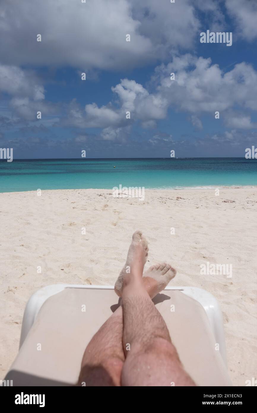 A man is laying on a beach chair with his feet up in the air. The beach is a beautiful blue color with the ocean in the background. The man is enjoyin Stock Photo