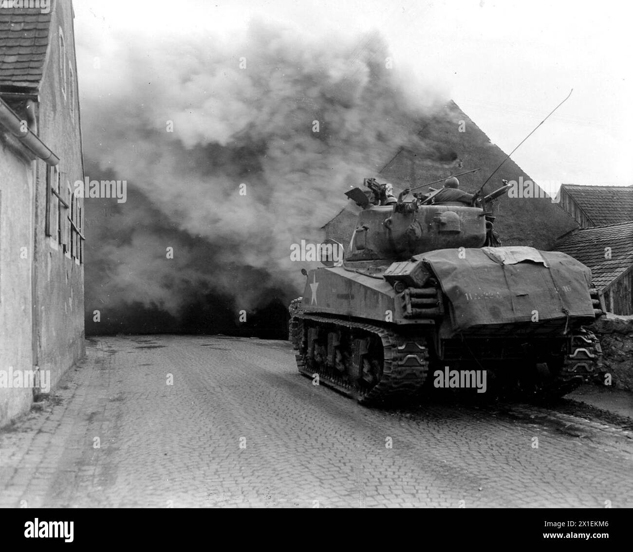 World War II Photos: Original Caption: Wernberg, Germany…Tank of 22nd Tank Battalion, 11th Armored Division, 3rd US Army, stands ready before the house its gun set on fire ca. April 1945 Stock Photo