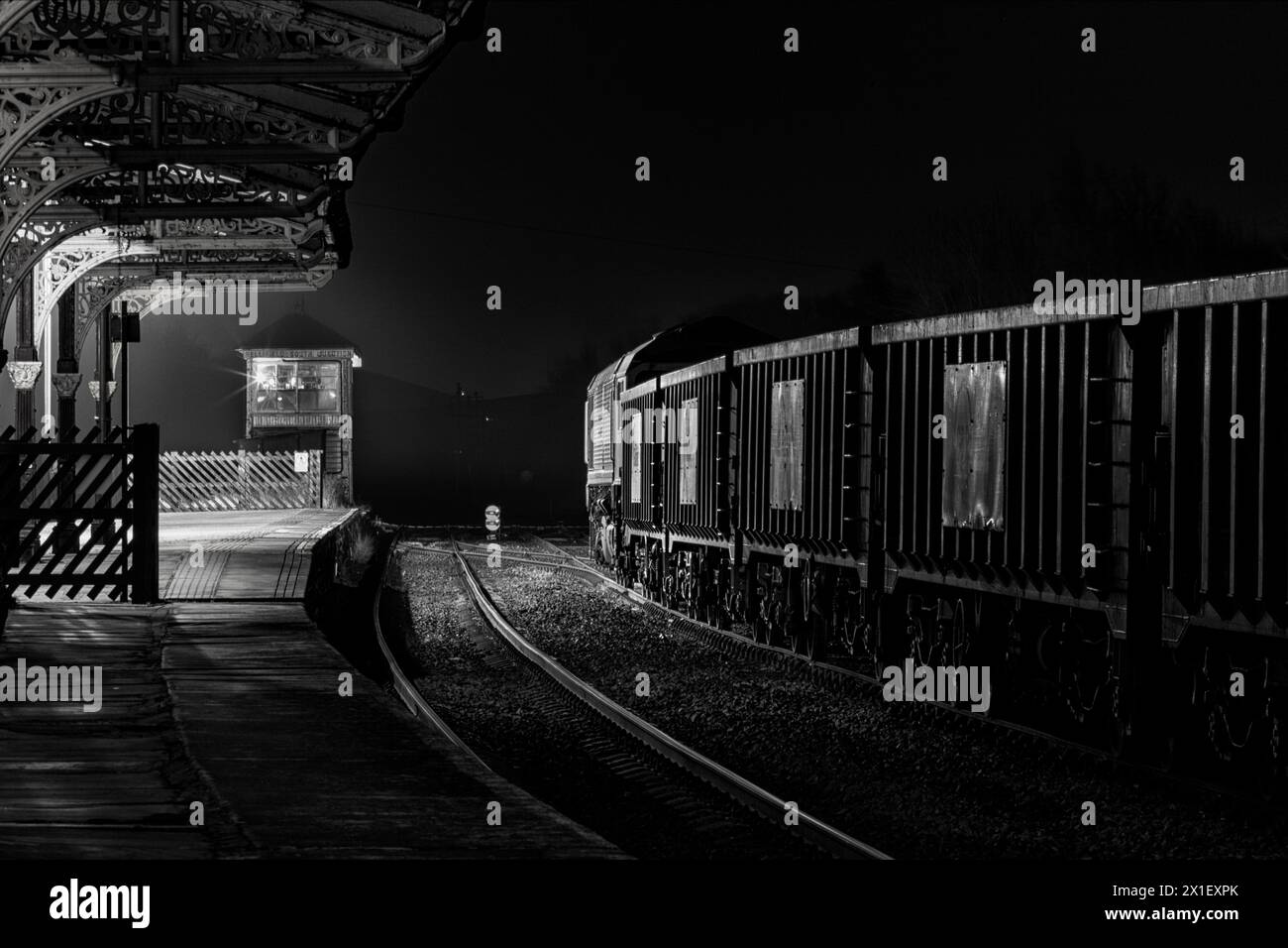GB Rail Freight class 66 locomotive 66718 under the Midland Railway canopy at  Hellifield  railway station with a train carrying aggregates at night Stock Photo
