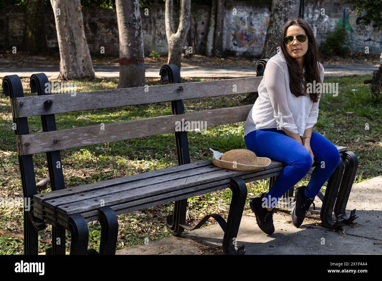 Young Latin American woman (33) sitting on a park bench with positive attitude for the arrival of spring. She is wearing blue pants, white shirt and g Stock Photo