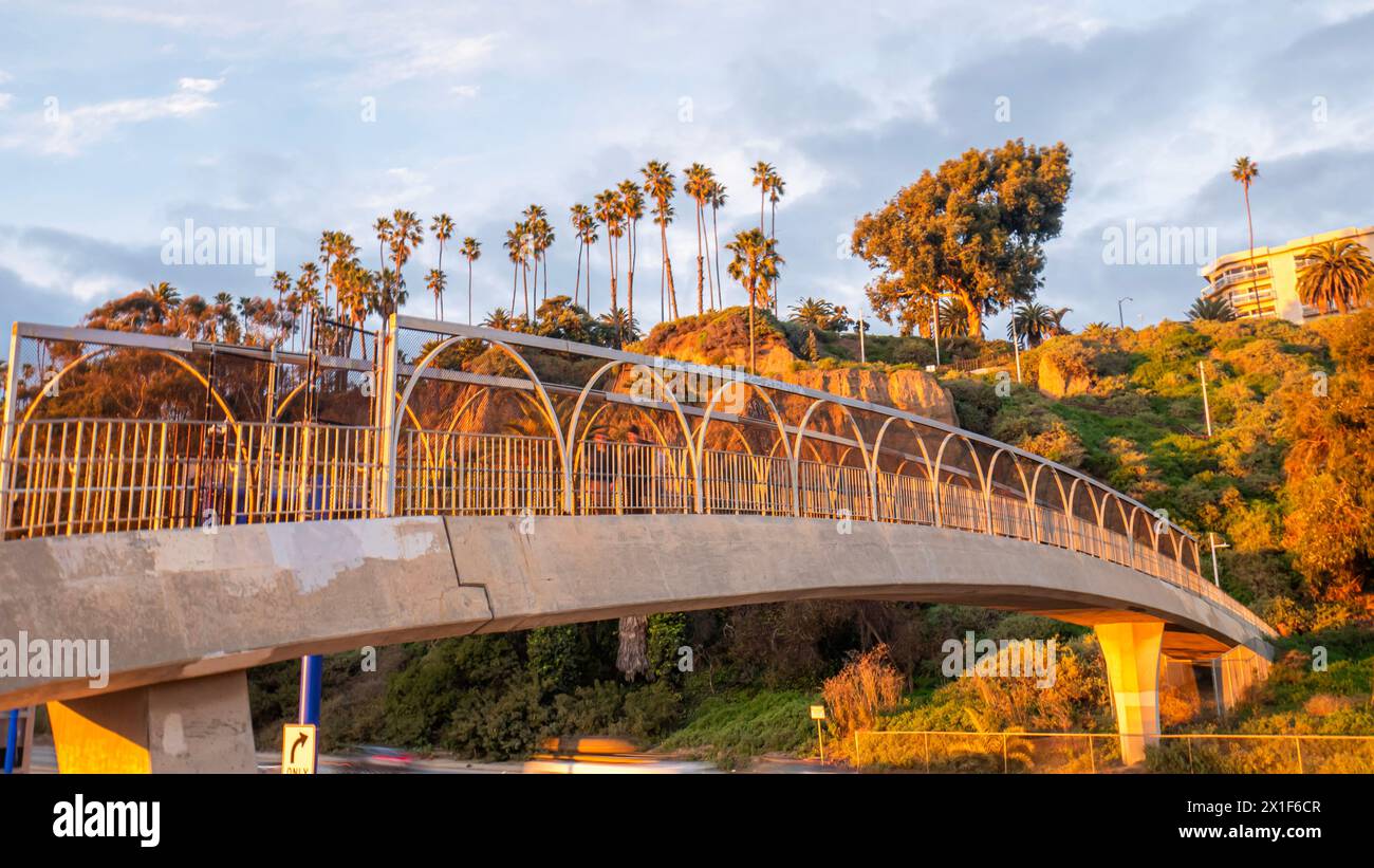 Sunset view of a concrete arched pedestrian bridge with railings, overlooking a scenic backdrop of palm trees and a hilly landscape, Santa Monica, CA Stock Photo