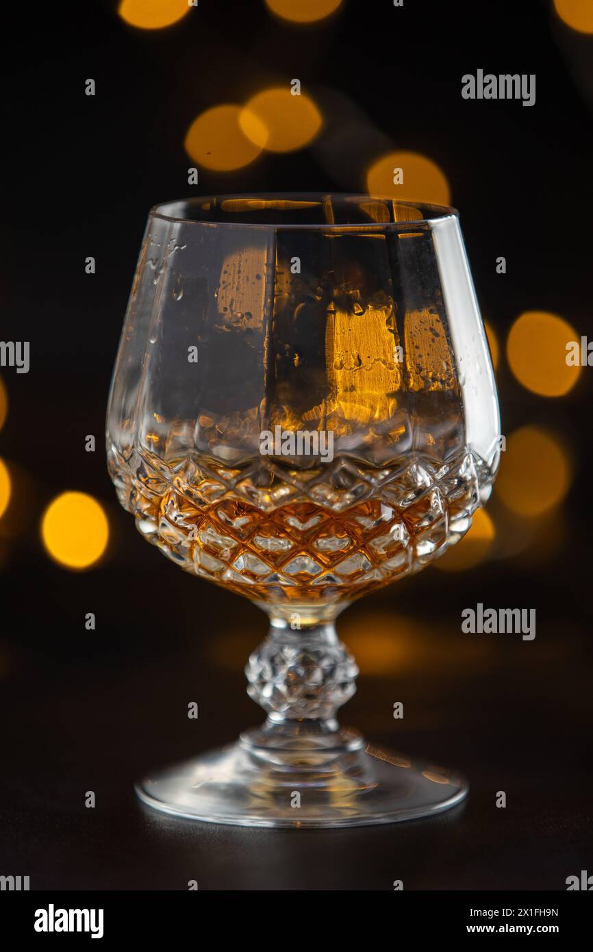 A glass of alcohol is sitting on a table. The glass is half full and has a yellowish tint. The image has a moody and mysterious feel to it, as the foc Stock Photo