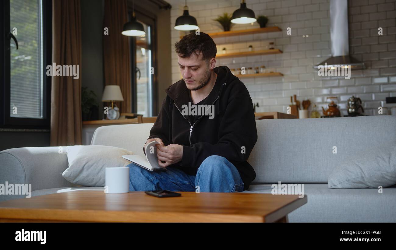 Man sitting on sofa takes the notebook from the table, looks through its pages, tears off a page and throws the notebook on the table at home. Stock Photo