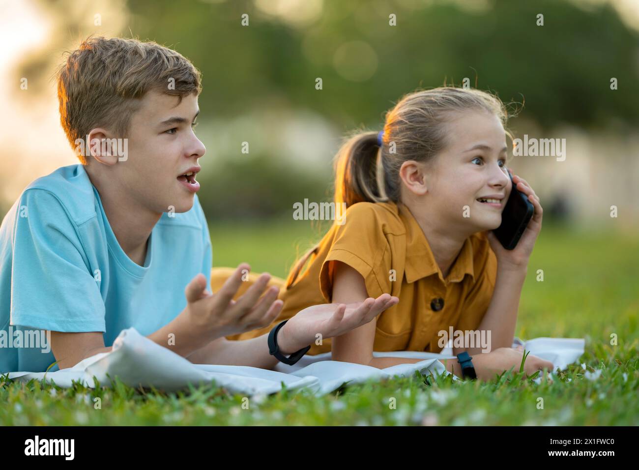 Teenage friends spending time outdoors while one is talking happily on mobile phone and another waiting frustrated at offensive behavior of her mate Stock Photo