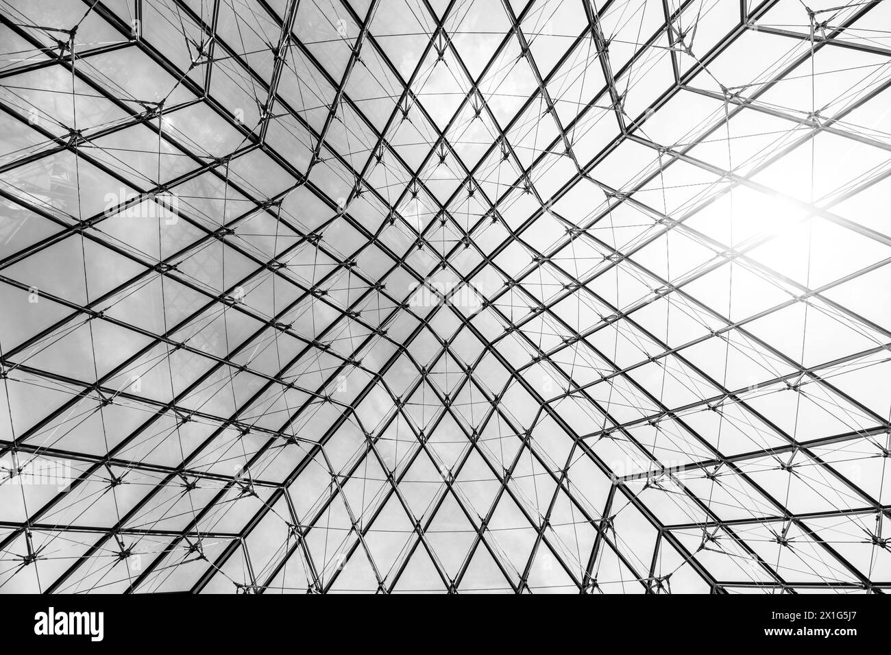 Underneath the Louvre Pyramid, the intricate glasswork contrasts against a blue sky on a clear day in Paris, France. Black and white image. Stock Photo