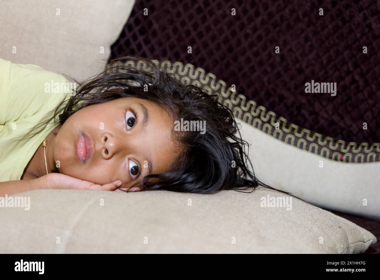 An image of a young girl lying on a couch in a living room, looking sad or disappointed. Stock Photo