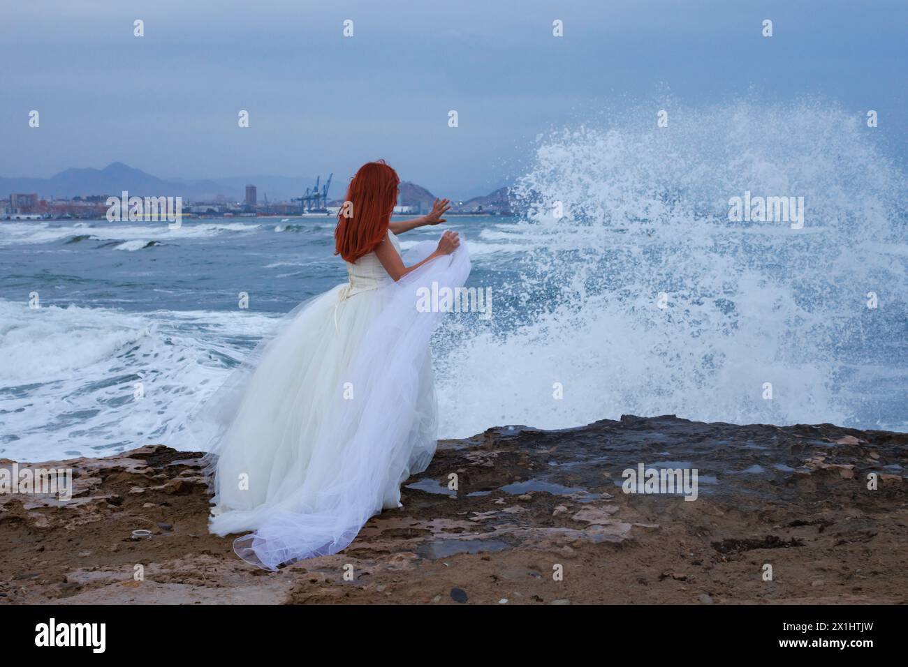 Redhead woman with wedding dress on the beach and splashing sea wave with stop splash gesture, Alicante, Spain Stock Photo