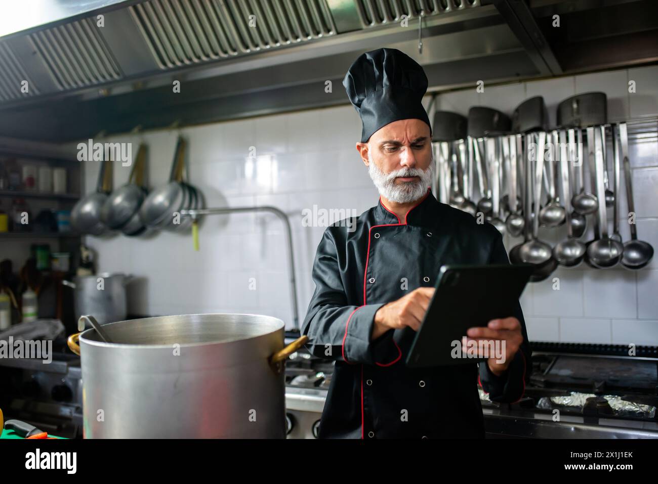 Seasoned male chef in uniform reads a digital recipe on a tablet while cooking Stock Photo