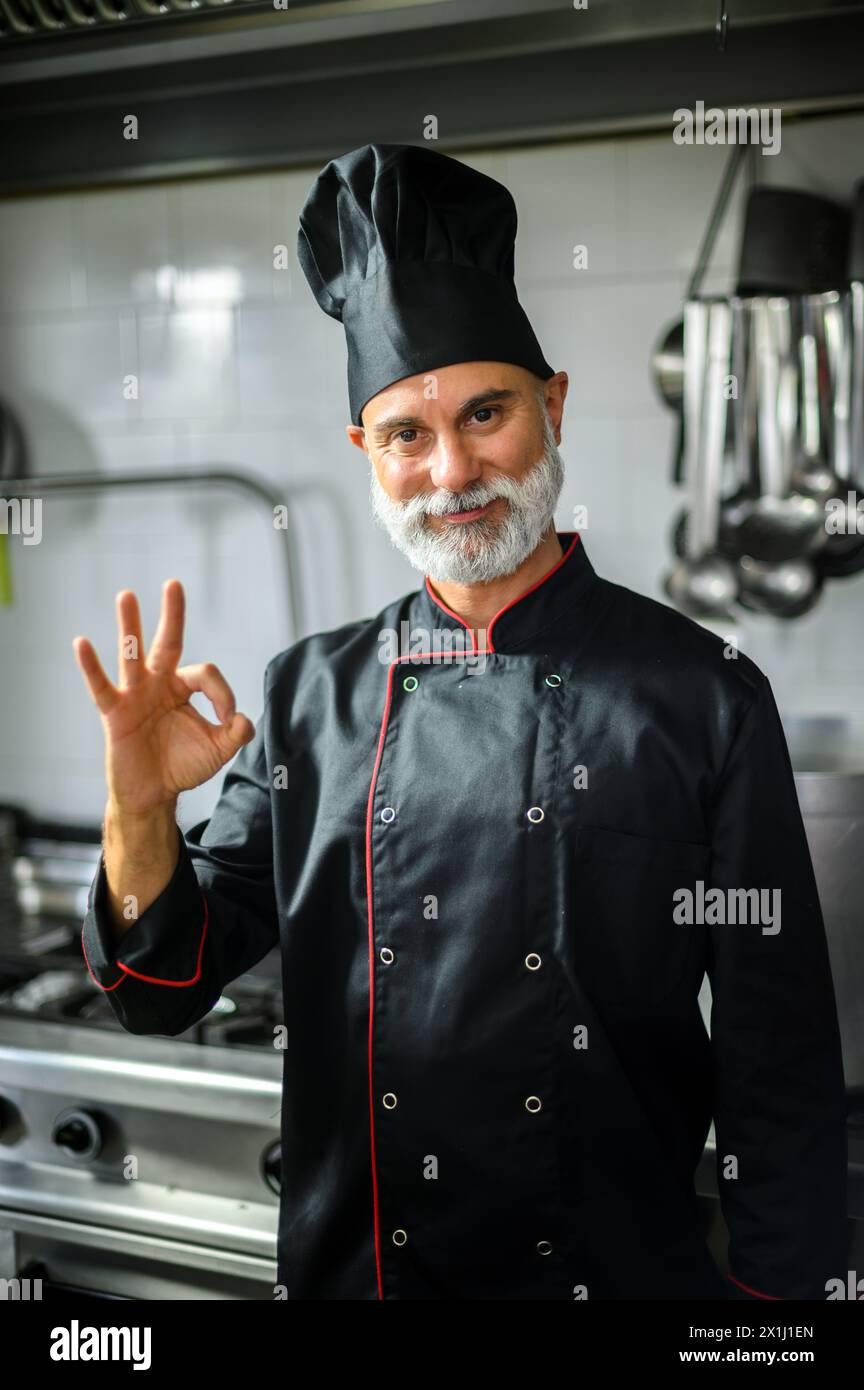 Professional chef wearing a black uniform gives a thumbs up in a commercial kitchen Stock Photo
