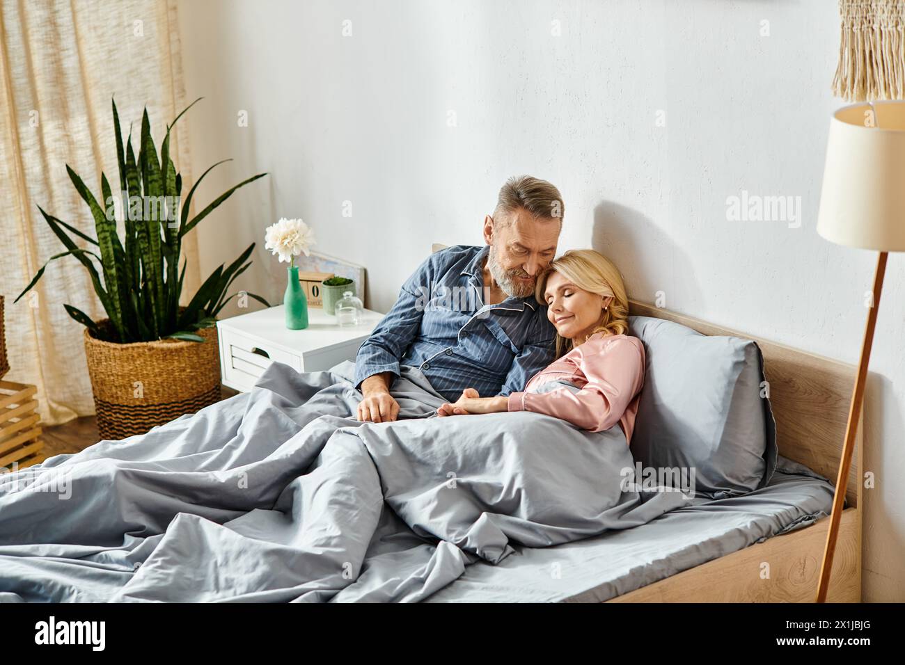 A man and woman, in cozy homewear, lovingly embrace while laying on a bed under a soft blanket in their bedroom. Stock Photo