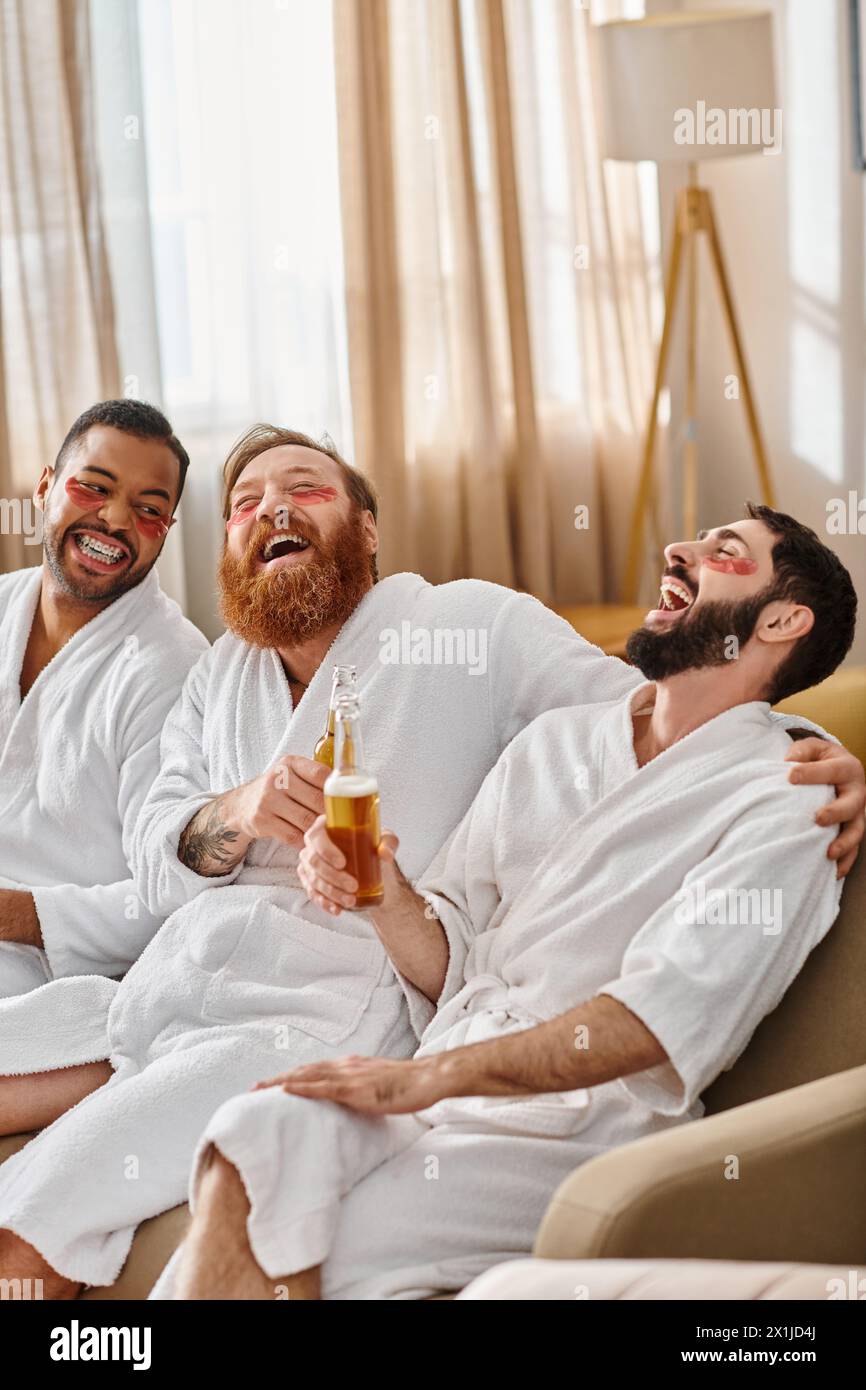 Three diverse, cheerful men in bathrobes having a great time sitting together on a lavish couch. Stock Photo