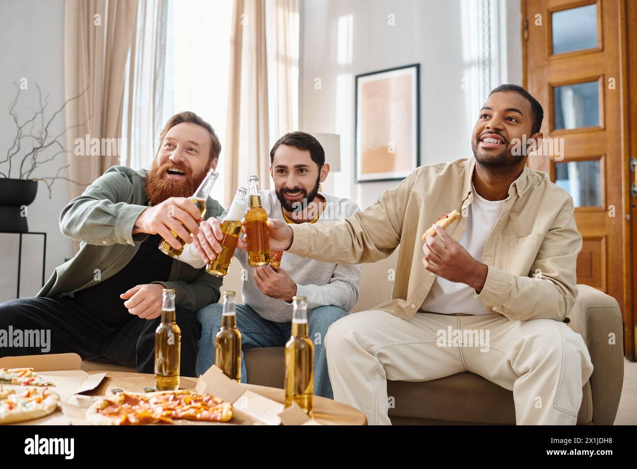 Three cheerful, handsome men of different races enjoy a casual get-together, sitting around a table with beers. Stock Photo