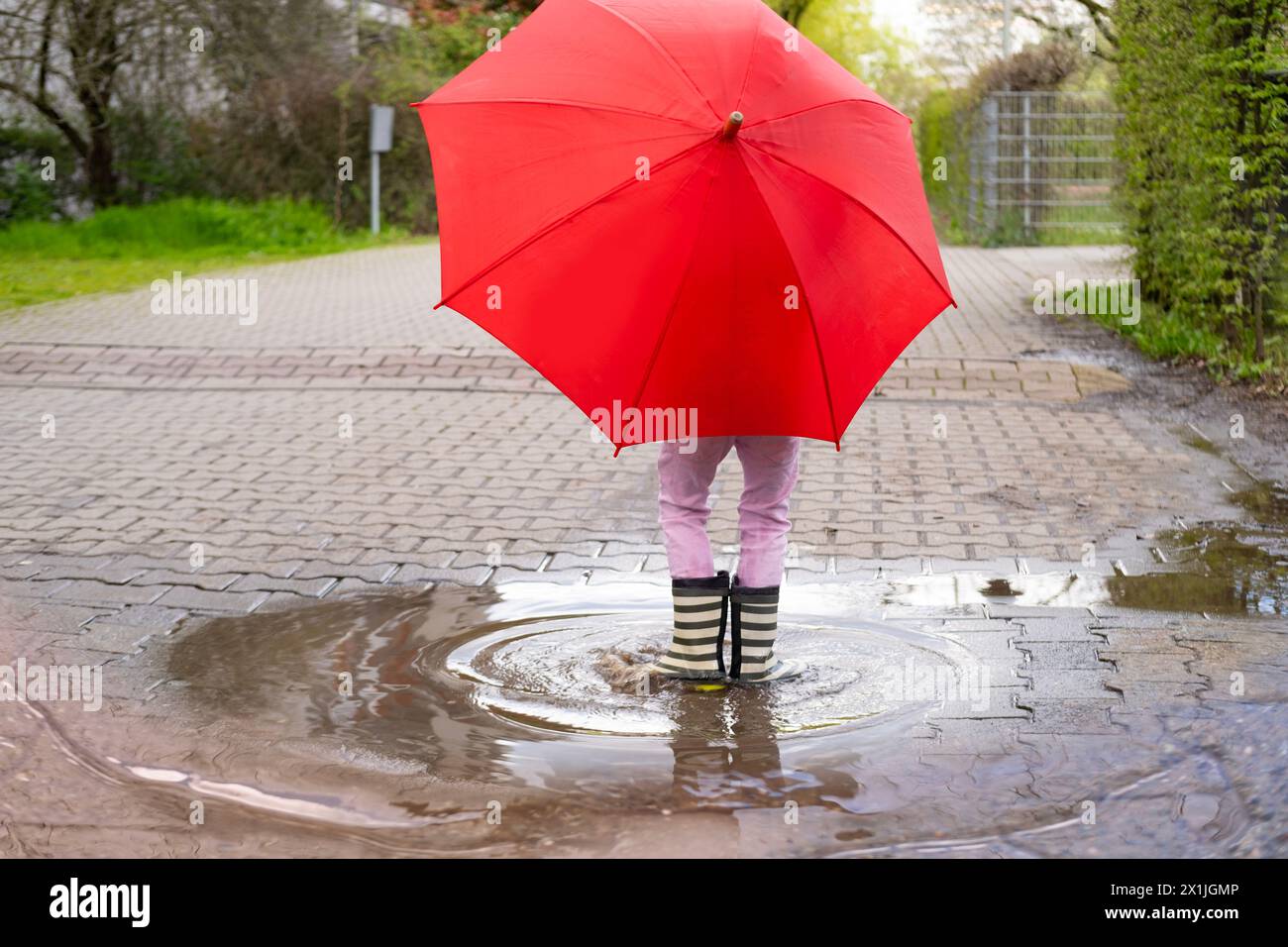 mischievous 5-year-old girl in rubber boots with red umbrella stands in rain puddle, capturing pure and simple joys childhood and magic rainy day Stock Photo