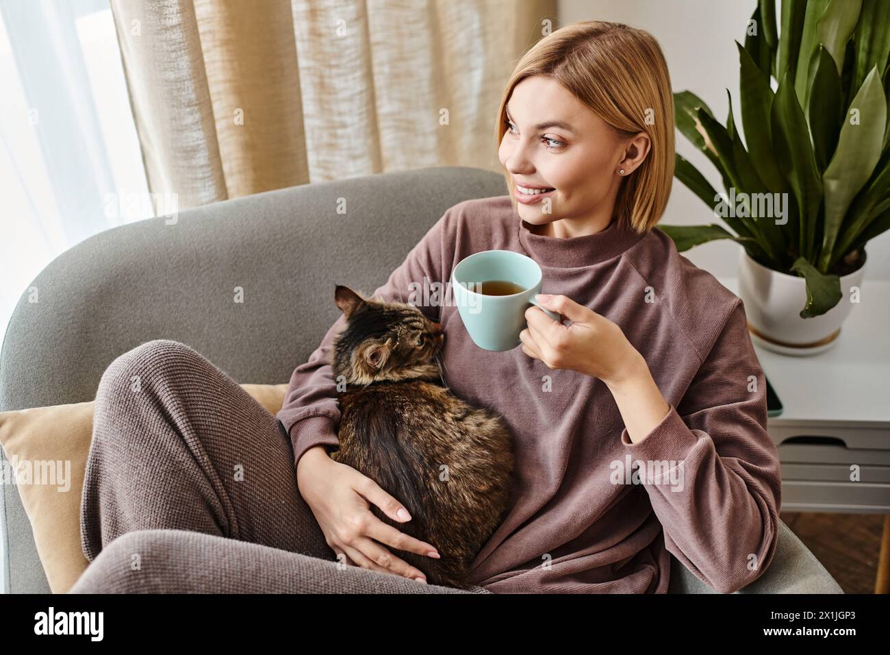 A woman with short hair sitting on a couch, cradling a cup of coffee while petting a cat on her lap. Stock Photo