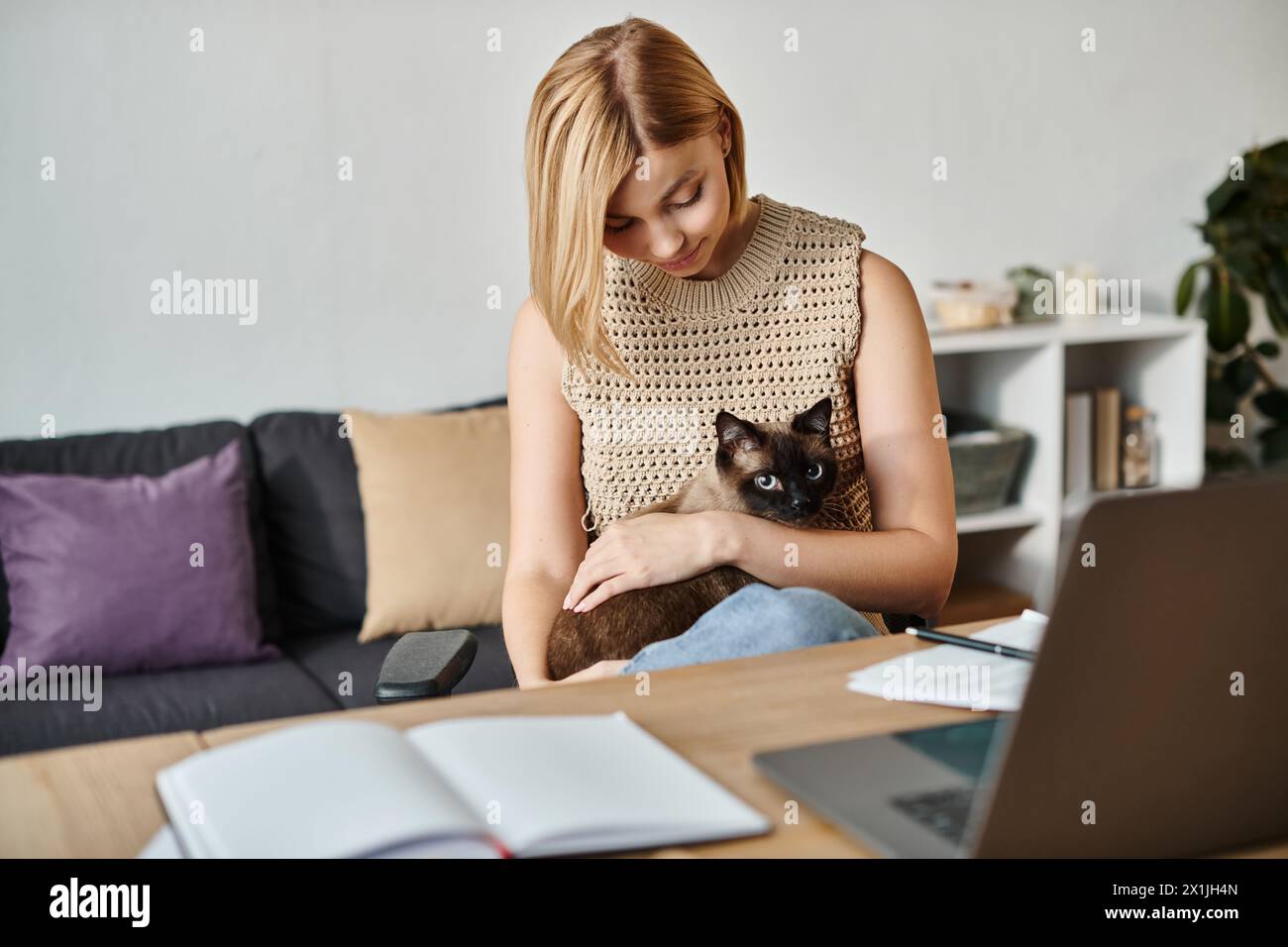 A contented woman with short hair sits at a table with her cat in her lap, enjoying a peaceful moment at home. Stock Photo