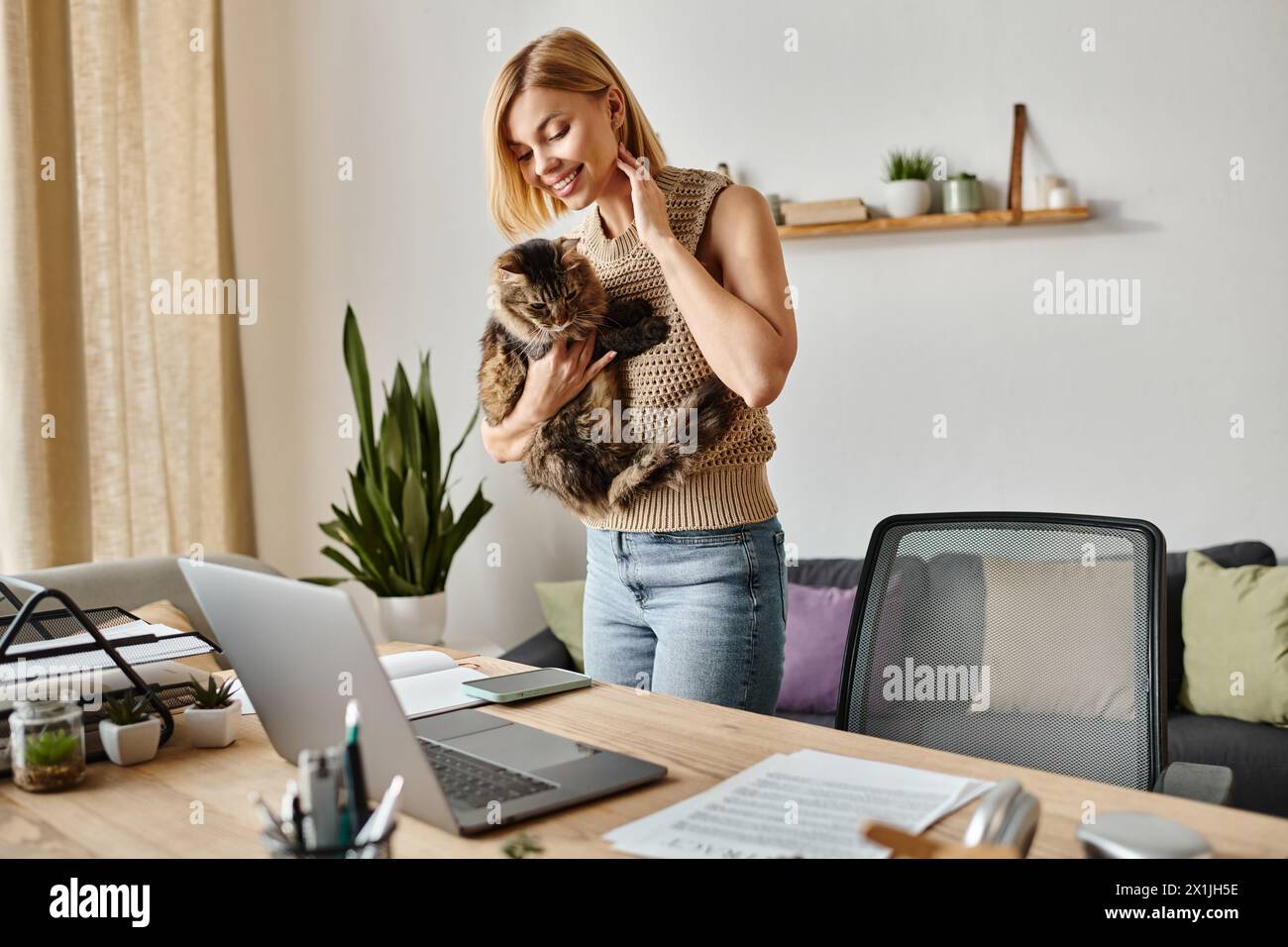 A woman holds her cat lovingly while standing by a laptop at home. Stock Photo